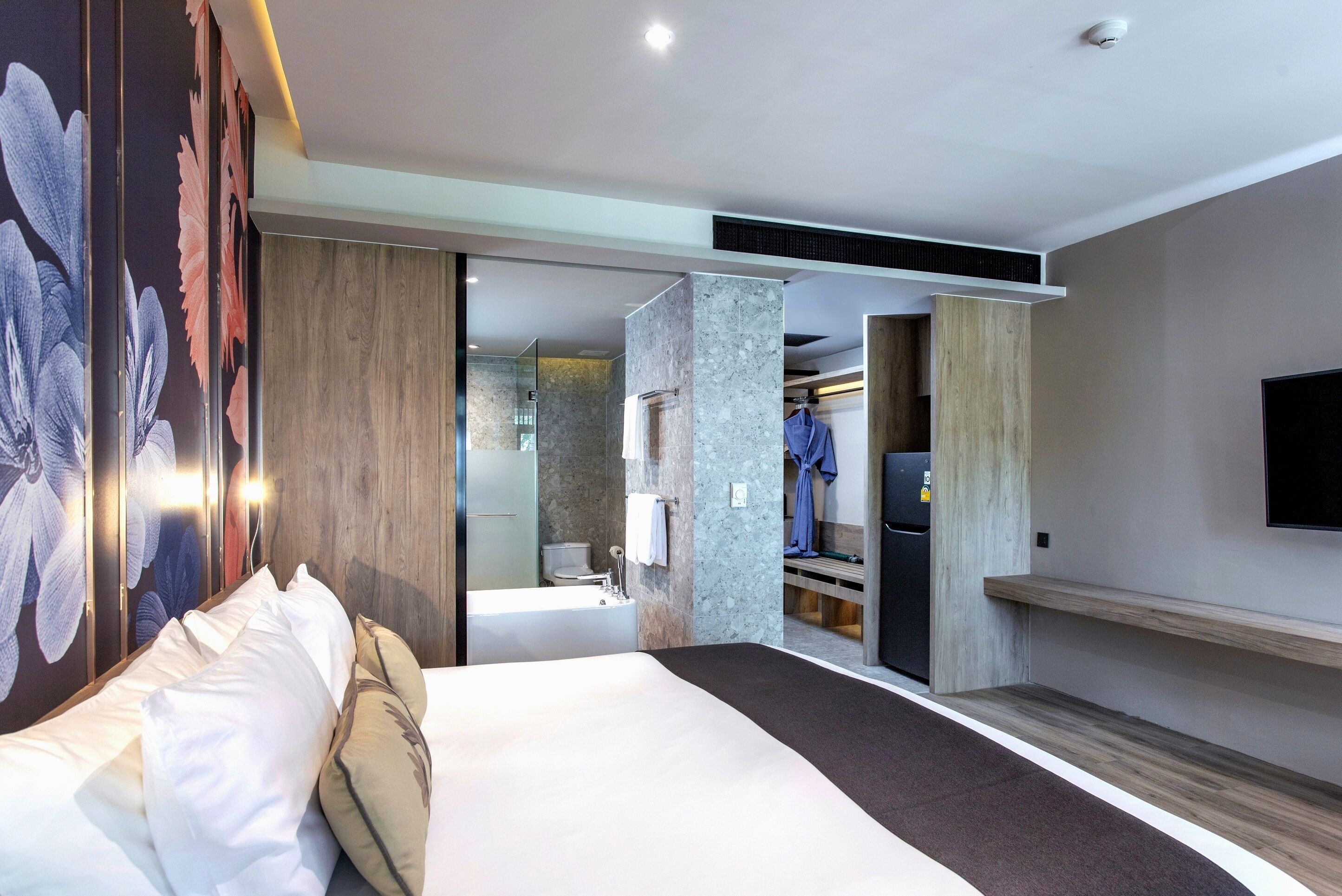 A modern hotel room with a large bed, a walk-in closet, a bathroom with a bathtub, and a flat-screen TV mounted on the wall.
