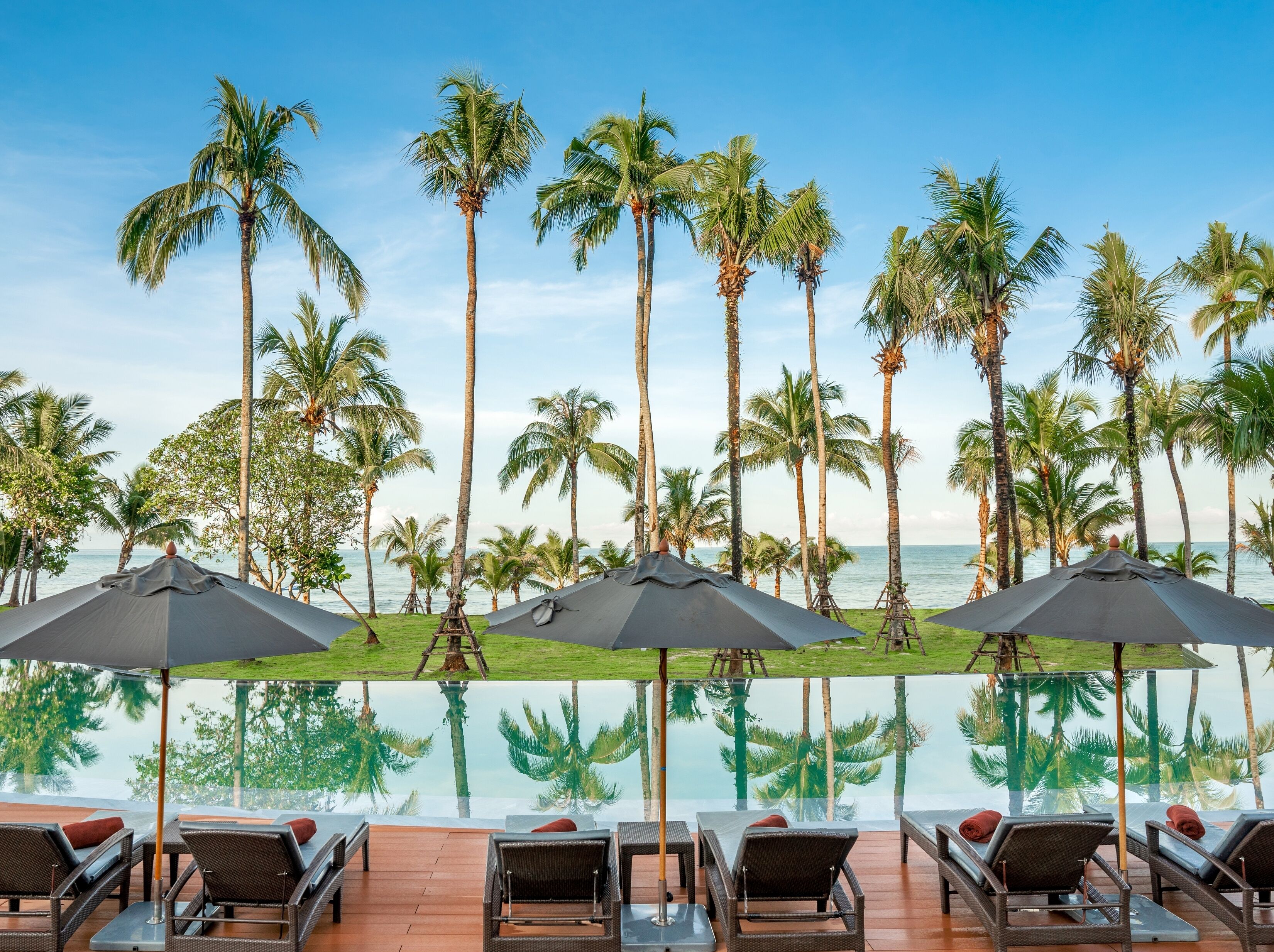 A poolside area with lounge chairs and umbrellas, surrounded by palm trees and overlooking a clear view of the ocean.