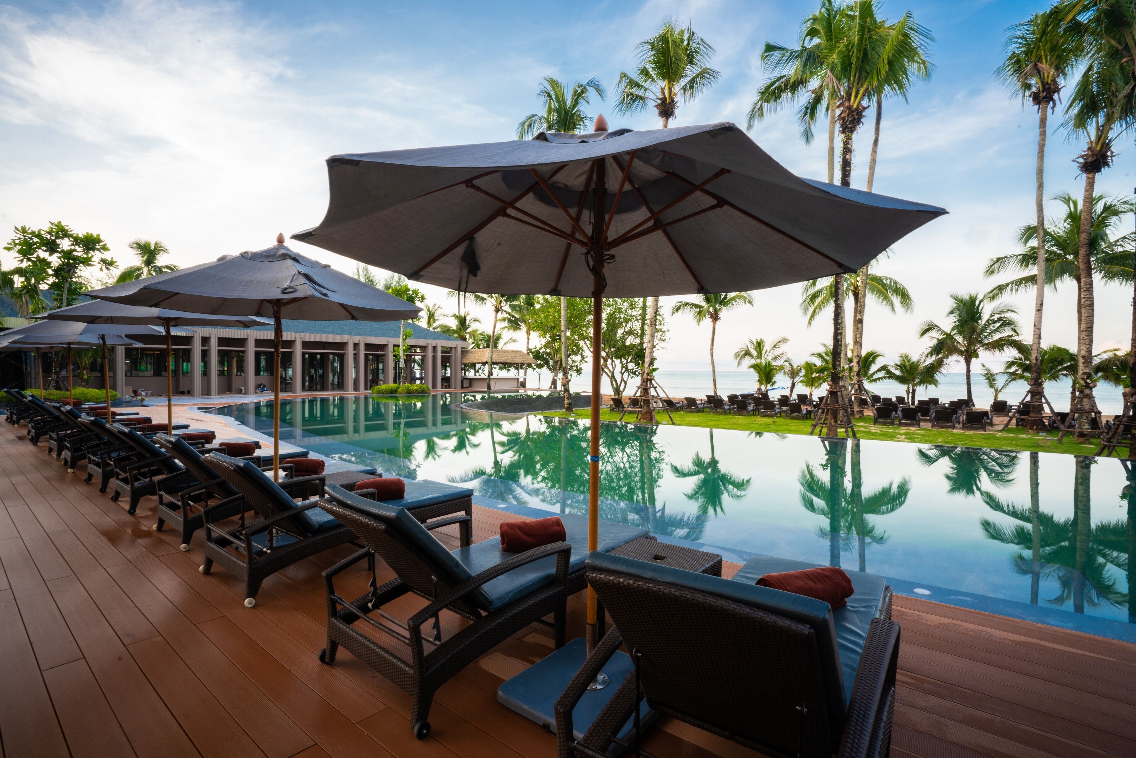A poolside area with lounge chairs and umbrellas, surrounded by palm trees and a clear view of the ocean.