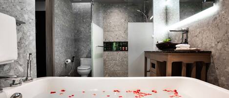 A modern bathroom with a marble wall, a bathtub with rose petals, a wooden vanity, and a glass shower enclosure.