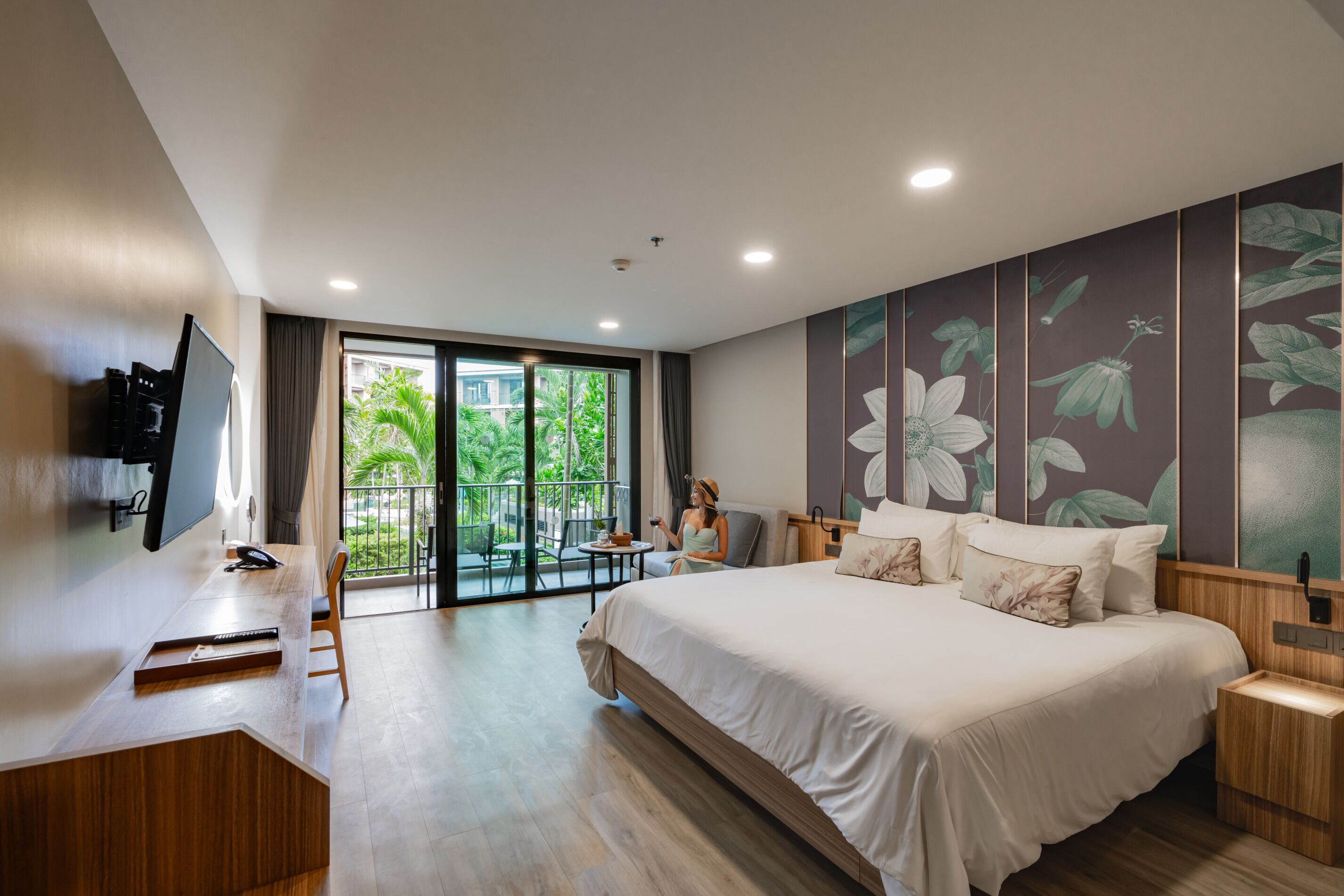 Habitación de hotel moderna con una cama grande, un escritorio, un televisor y un balcón con vistas a zonas verdes.