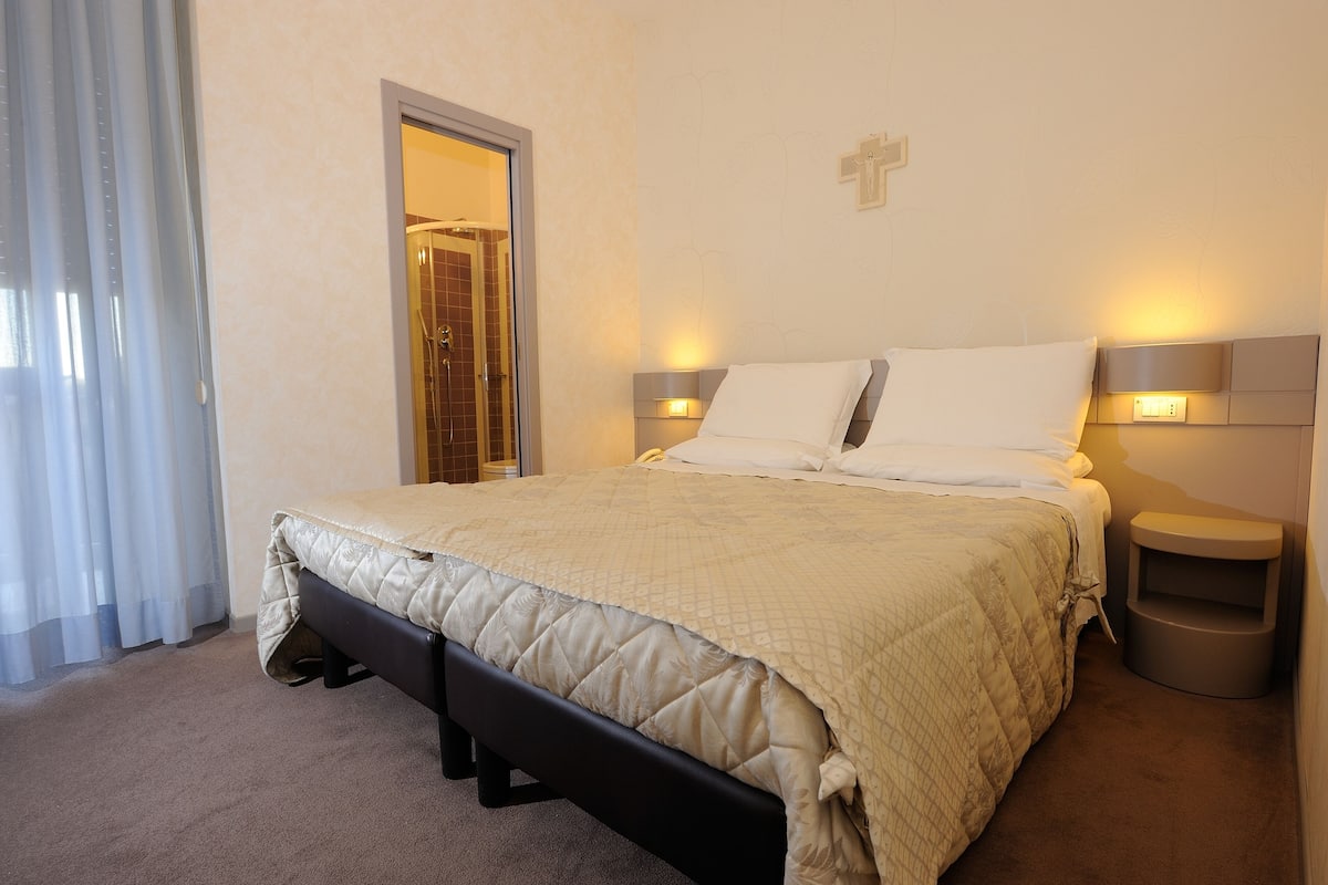 Habitación de hotel con cama, mesitas de noche, un banco, ducha y una cruz en la pared.