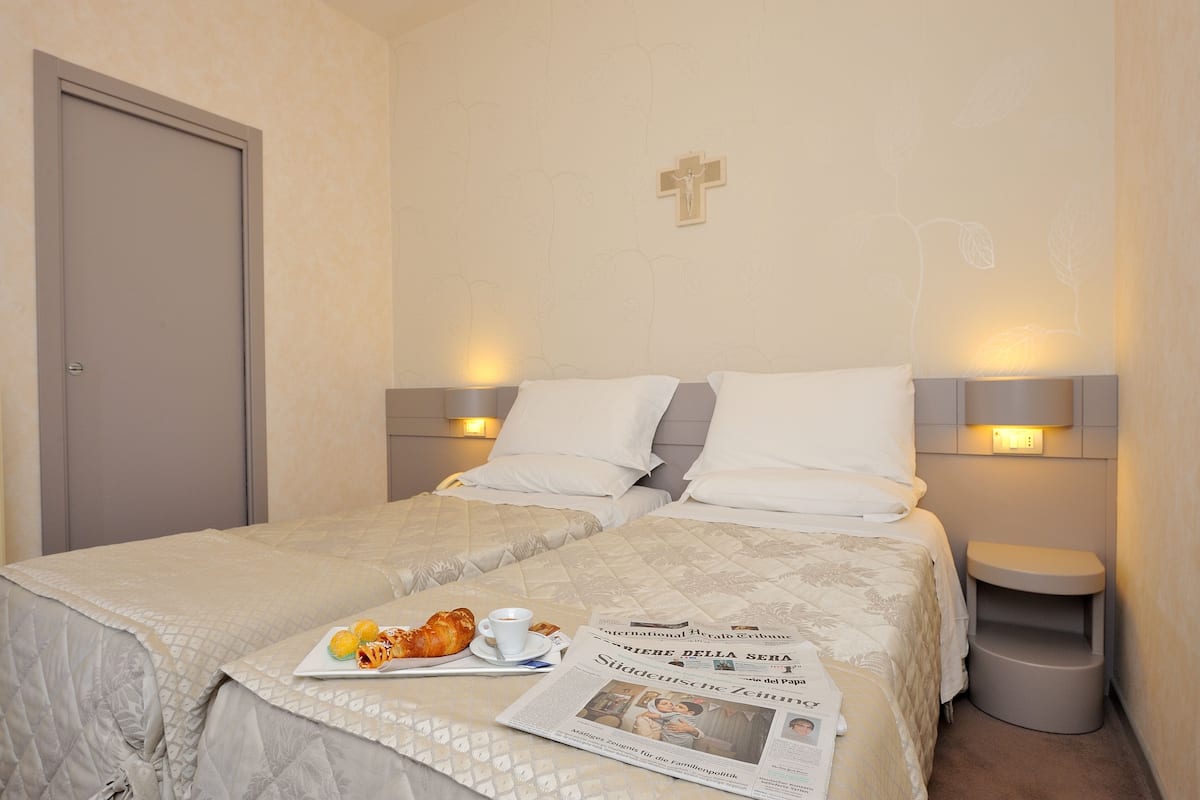 Habitación de hotel con cama, lámparas de noche, mesita auxiliar, un cubo de basura y una bandeja de desayuno con croissants y un periódico.