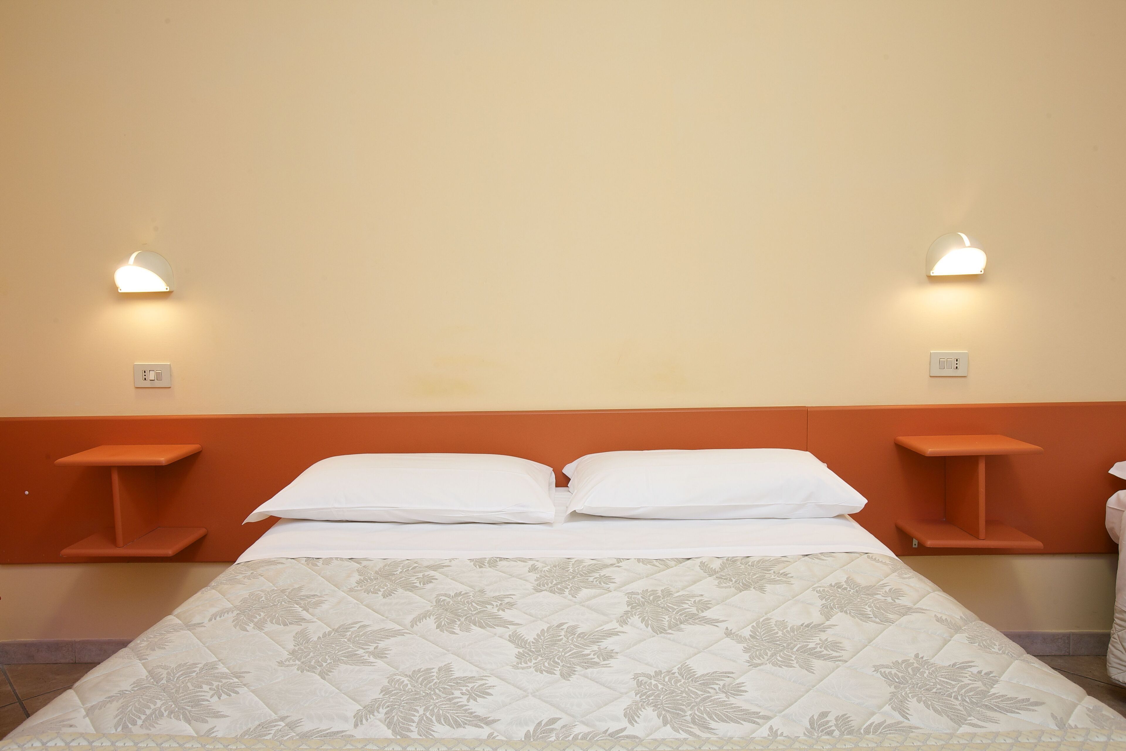 Un lit bien fait, recouvert d’une couvre-lit à motifs floraux, deux tables de chevet avec des lampes, et un mur sobre avec un interrupteur.