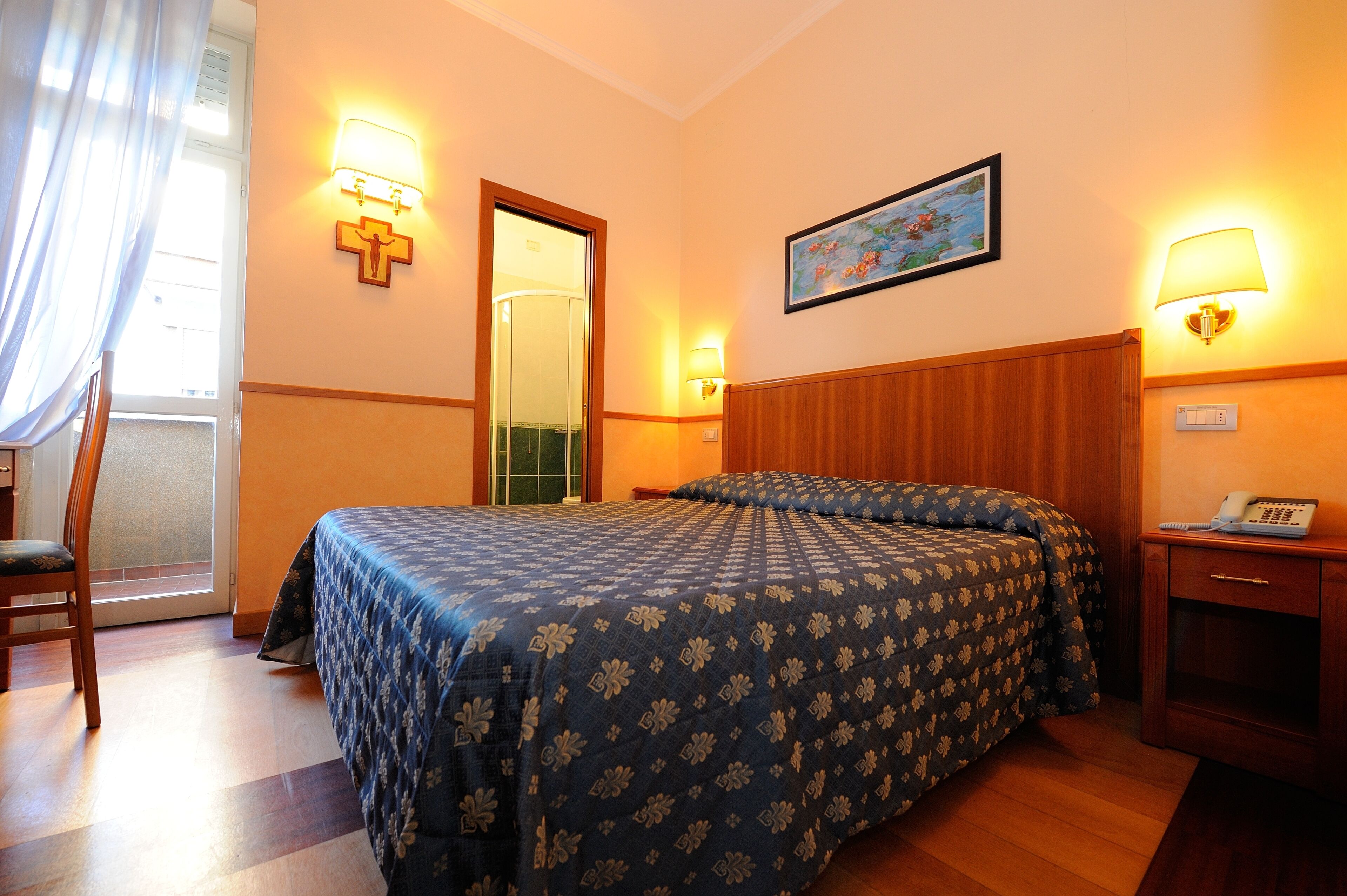 Quarto de hotel com cama, uma cadeira, escrivaninha com telefone e um quadro na parede.