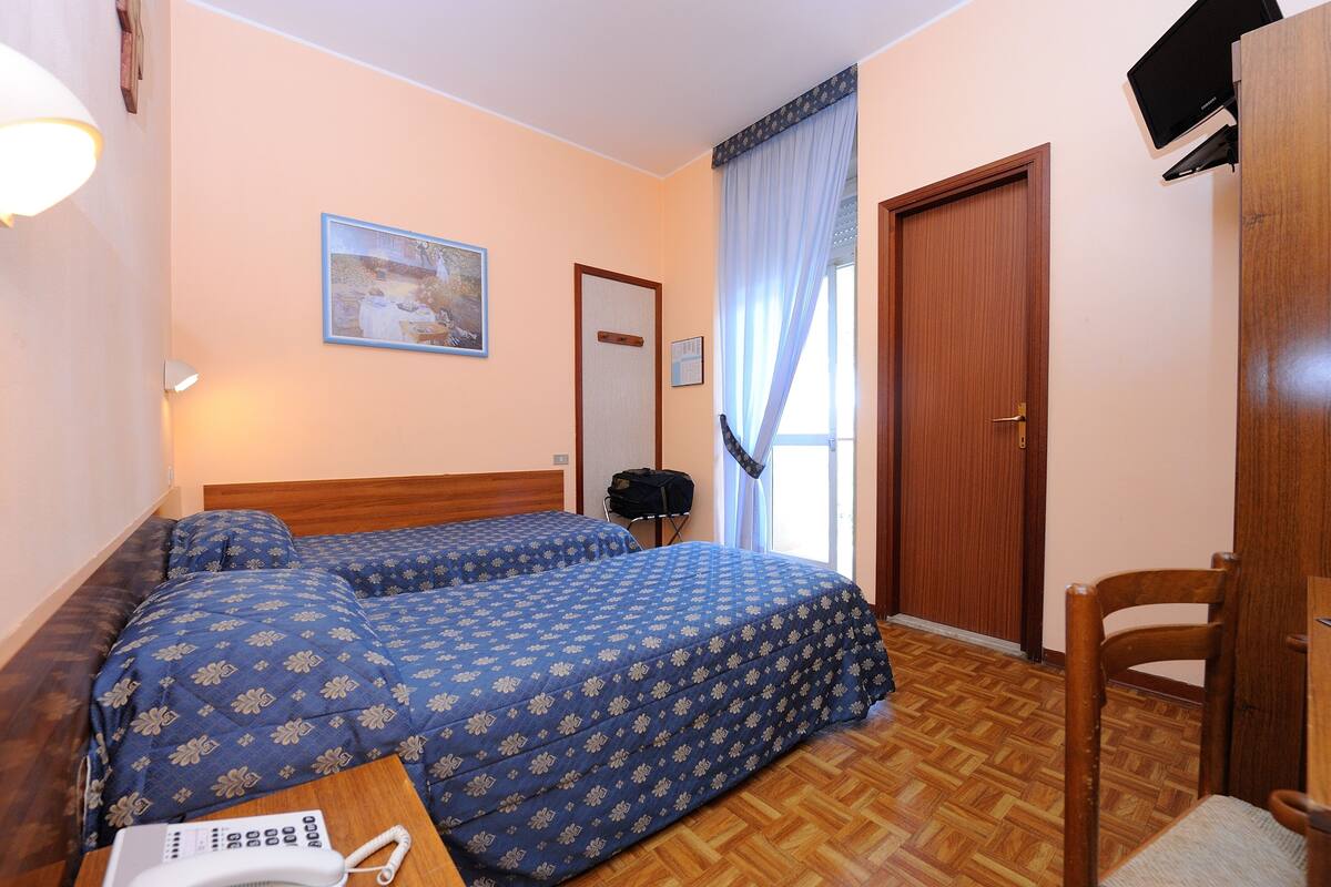 Habitación de hotel con dos camas, cabecero de madera, una mesita con teléfono, una silla, televisor montado en la pared y ventana con cortinas.