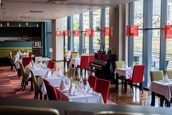 A restaurant with tables set for dining, a piano, and large windows with red blinds.