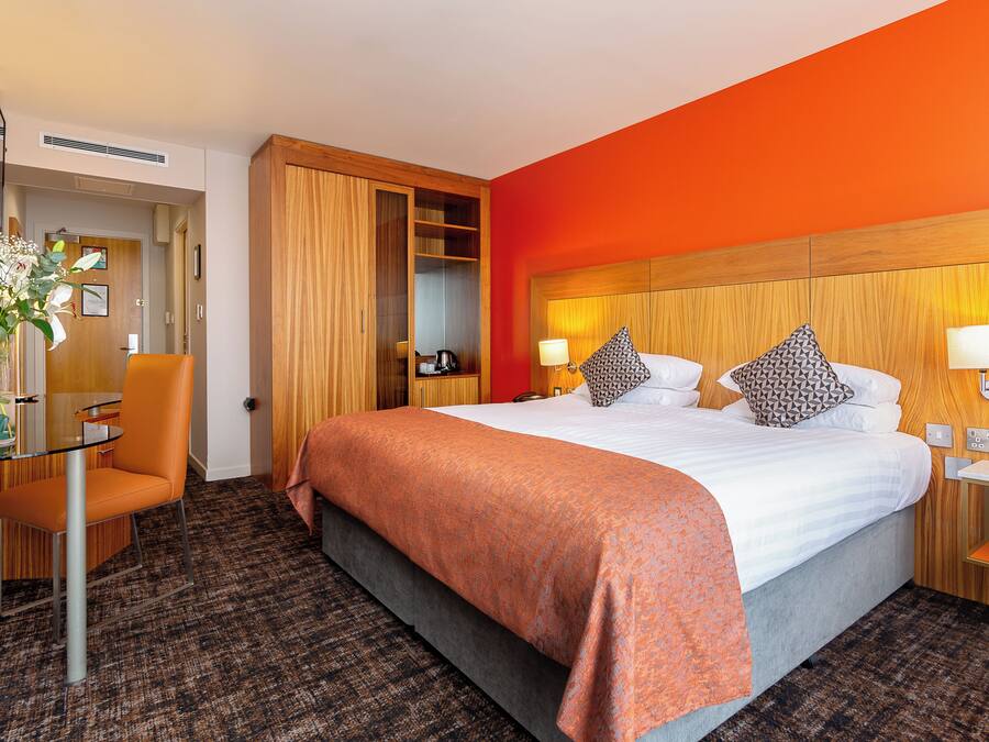 Habitación de hotel con una cama grande, un armario de madera, un escritorio con silla y una mesita con lámpara.