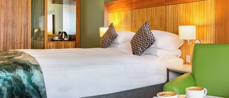 A hotel room with a large bed, two pillows, a bedside table with a lamp, a green armchair, and a small table with two cups of coffee and a plate of food.