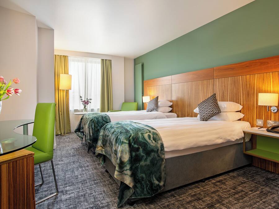 A hotel room with two beds, a green chair, a glass table with a vase of flowers, and a wooden headboard.