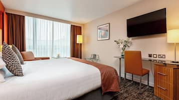 Quarto de hotel com uma cama grande, uma escrivaninha, uma cadeira, uma televisão e uma janela com cortinas.