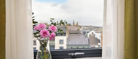 A window with a vase of pink flowers on a window sill, overlooking a cityscape with buildings and a church spire.