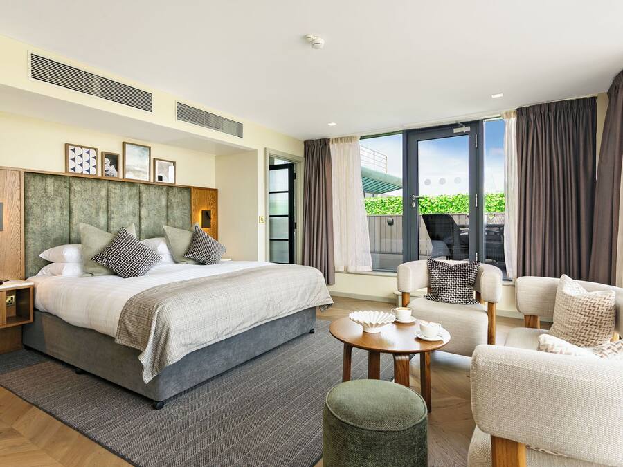 A modern hotel room with a large bed, a seating area with a table, and a balcony with outdoor furniture.