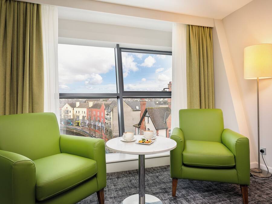 Habitación de hotel con dos sillones verdes, una pequeña mesa redonda con una tetera y tazas, y una lámpara de pie.