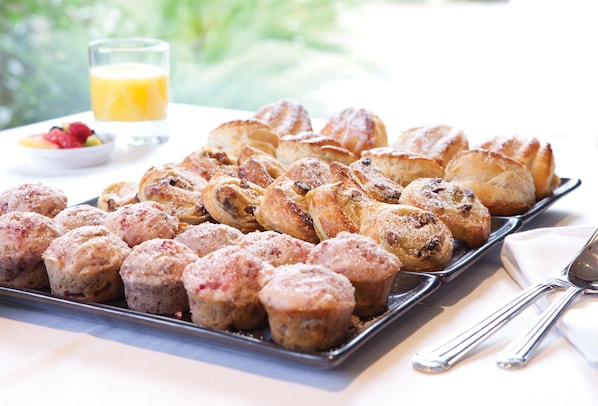 Una bandeja con pasteles y muffins sobre una mesa, junto con un vaso de jugo de naranja y un bol de fruta.