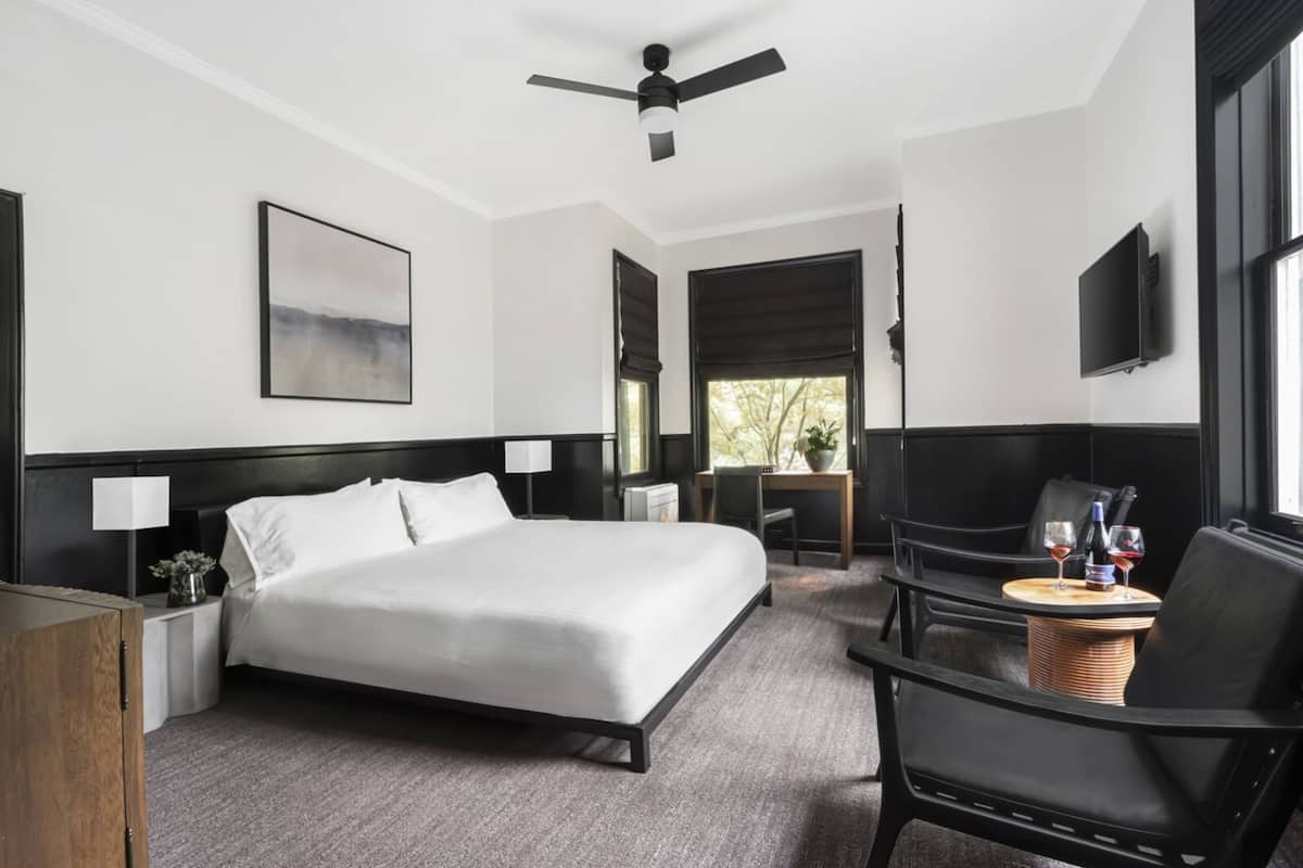 A hotel room with a bed, bedside tables, a desk, a chair, a television, and a window with a view of trees.