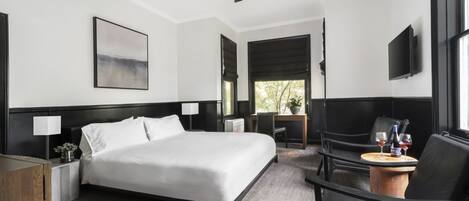 Habitación de hotel con cama, mesitas de noche, escritorio, silla, televisor y ventana con vista a árboles.