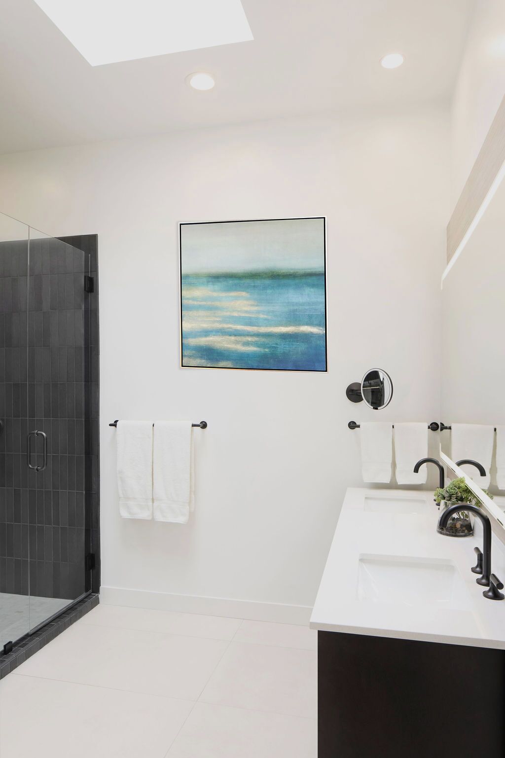 A modern bathroom with a glass shower enclosure, a white sink with a black faucet, and a framed painting of a seascape on the wall.