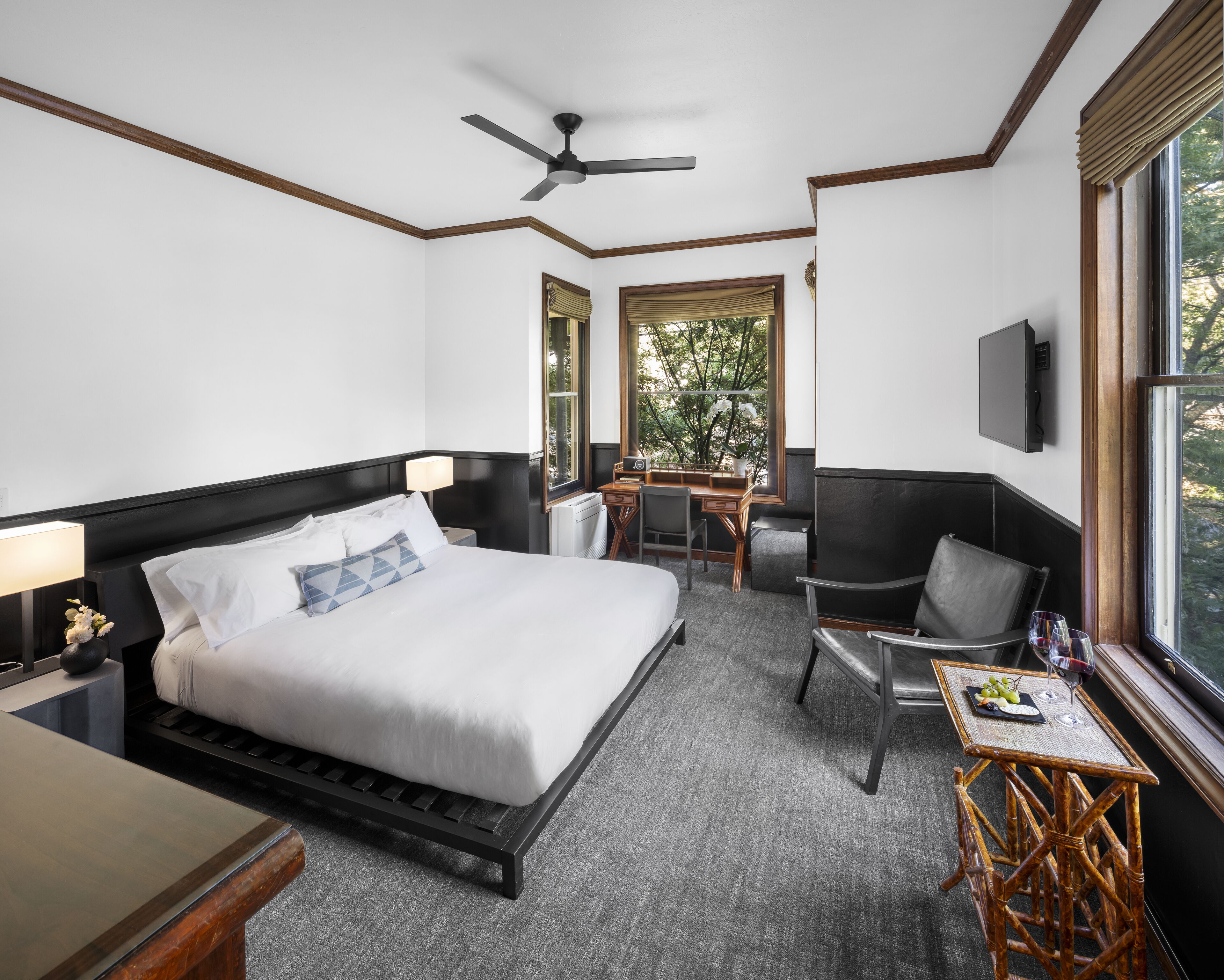A hotel room with a bed, a desk, a chair, a small table, a TV, and a window with a view of trees.