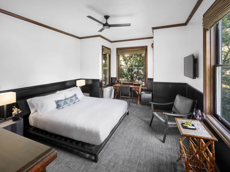 A hotel room with a bed, a desk, a chair, a small table, a TV, and a window with a view of trees.