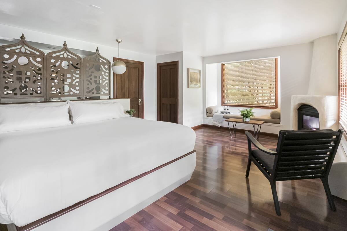 A spacious bedroom with a large bed, a wooden floor, a seating area with a bench and a small table, and a fireplace.