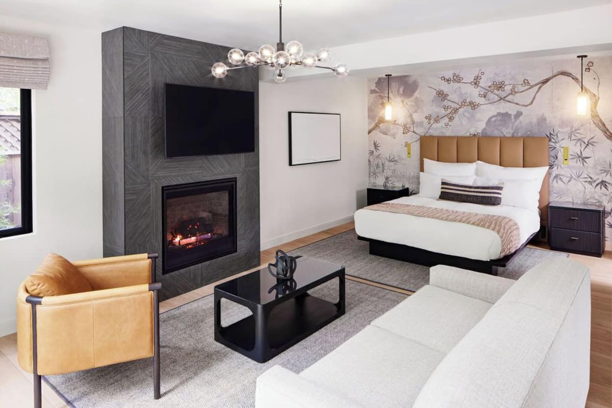 A modern bedroom with a fireplace, a bed with a headboard, a sofa, a coffee table, and a TV mounted on the wall.