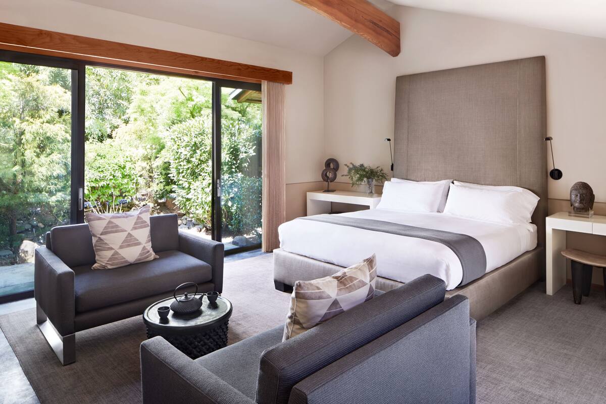 A modern bedroom with a large bed, two sofas, a small table, and a view of greenery through large windows.