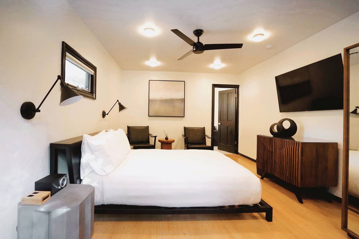 A modern hotel room with a large bed, a ceiling fan, a TV, and a small table with chairs.