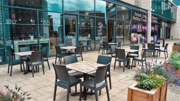 An outdoor seating area with tables and chairs, a bar, and a glass facade with signage for events. - Holiday Inn Express Leeds City Centre Armouries by IHG (Leeds)