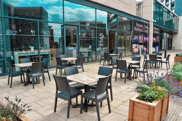 An outdoor seating area with tables and chairs, a bar, and a glass facade with signage for events. - Holiday Inn Express Leeds City Centre Armouries by IHG (Leeds)