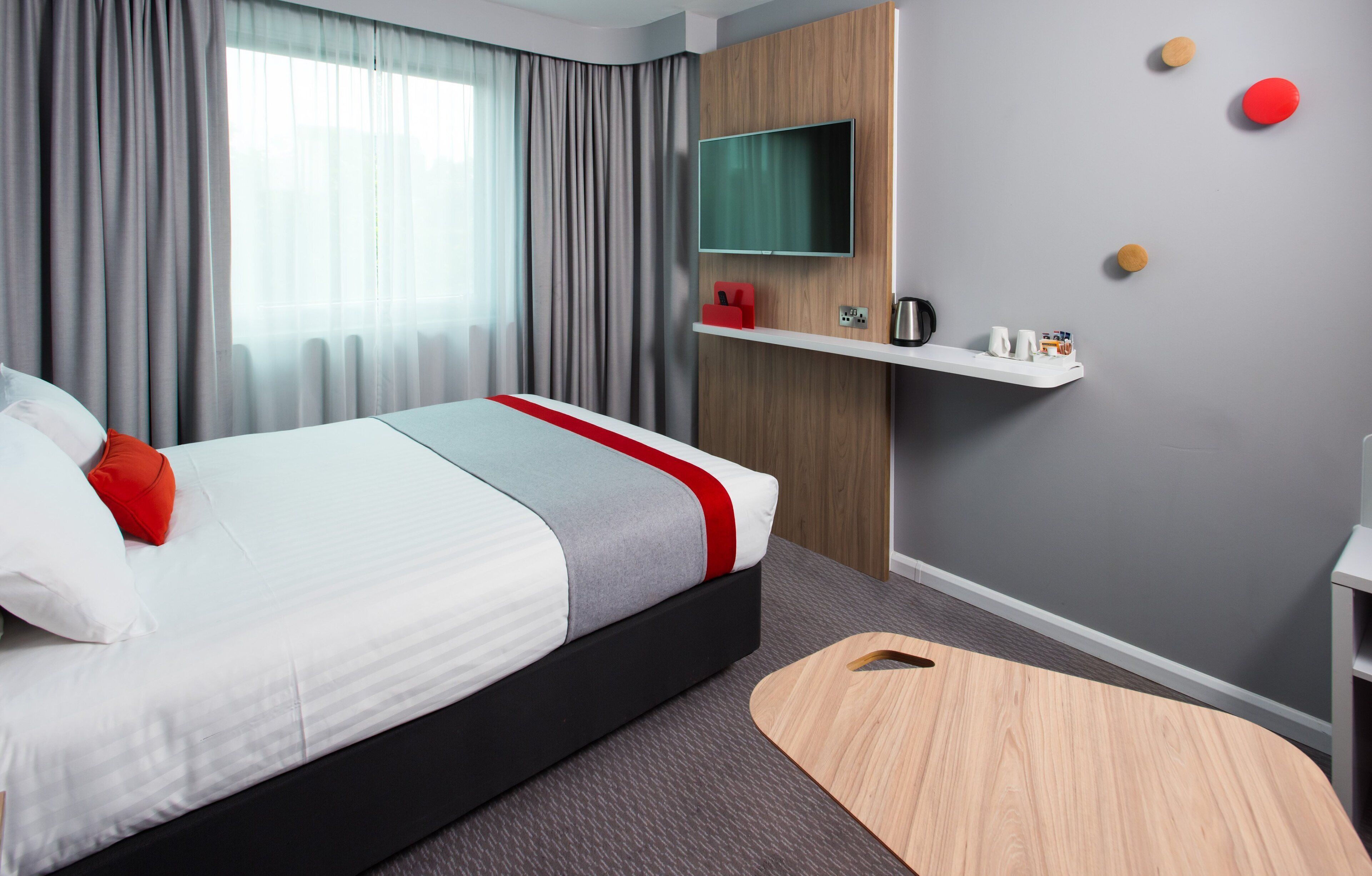 A modern hotel room with a bed, a TV mounted on the wall, a small shelf with toiletries, and a wooden side table.