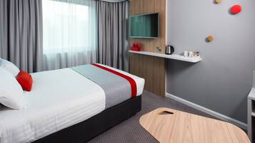 A modern hotel room with a bed, a TV mounted on the wall, a small shelf with toiletries, and a wooden side table.