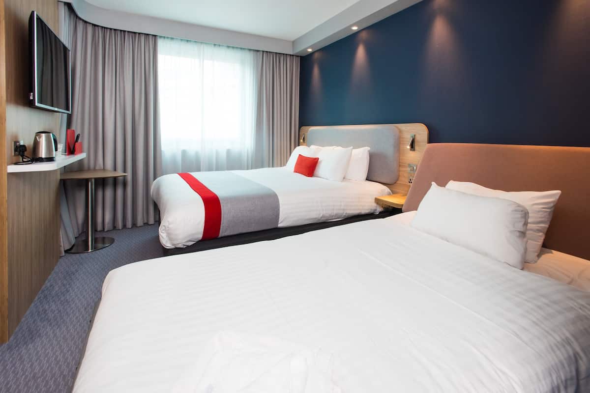 A hotel room with two beds, a TV, a desk with a kettle, and a view of the outdoors.