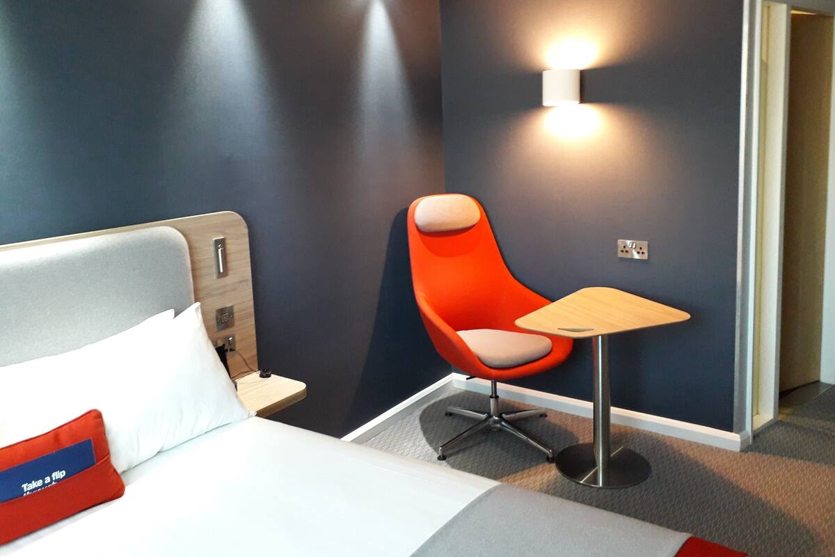A modern hotel room with a bed, a bright orange chair, a small table, and a wall-mounted light.