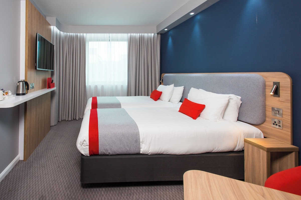 A modern hotel room with a large bed, a TV mounted on the wall, a bedside table, and a window with curtains.