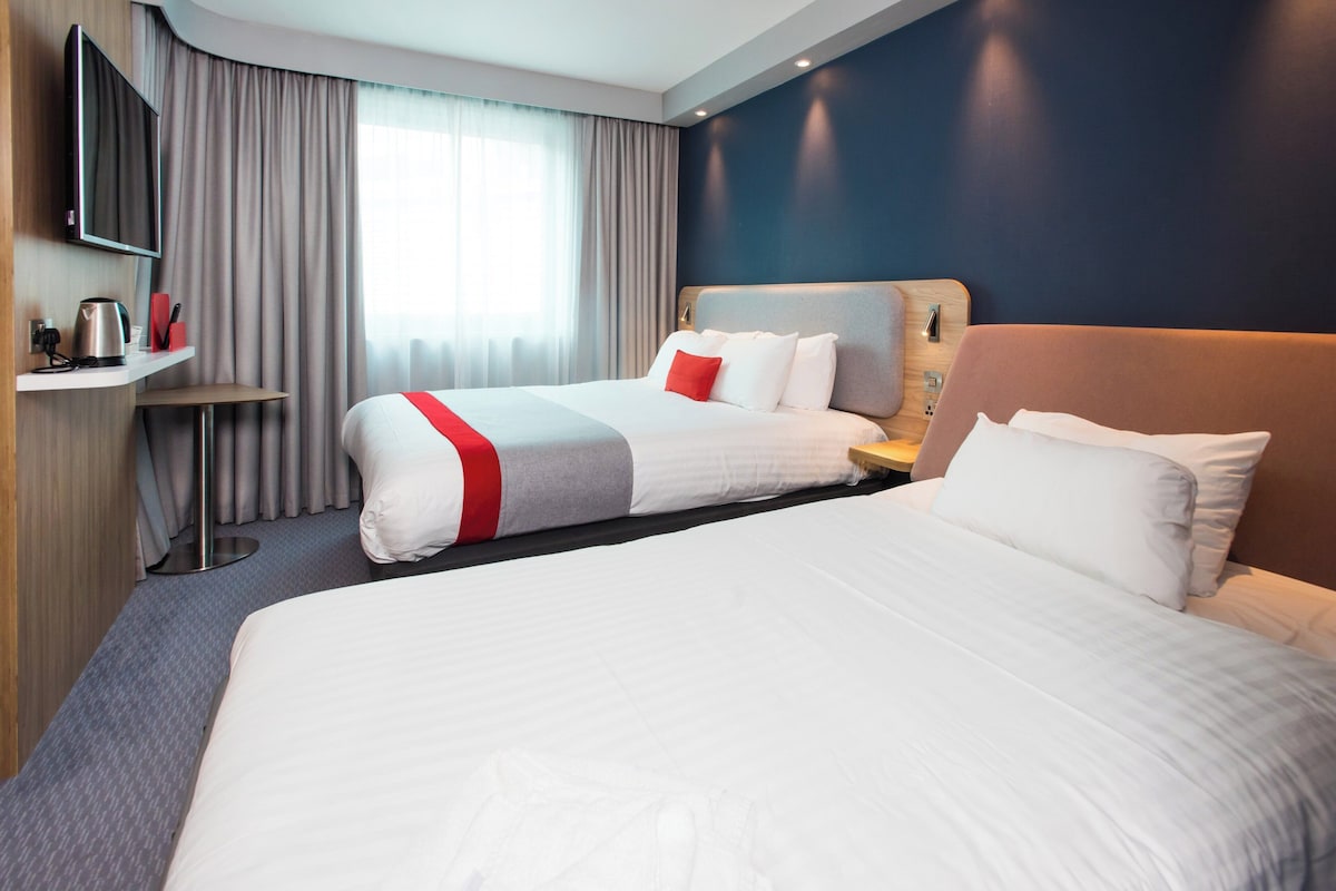 A hotel room with two beds, a TV, a desk with a kettle, and a view of the outdoors.