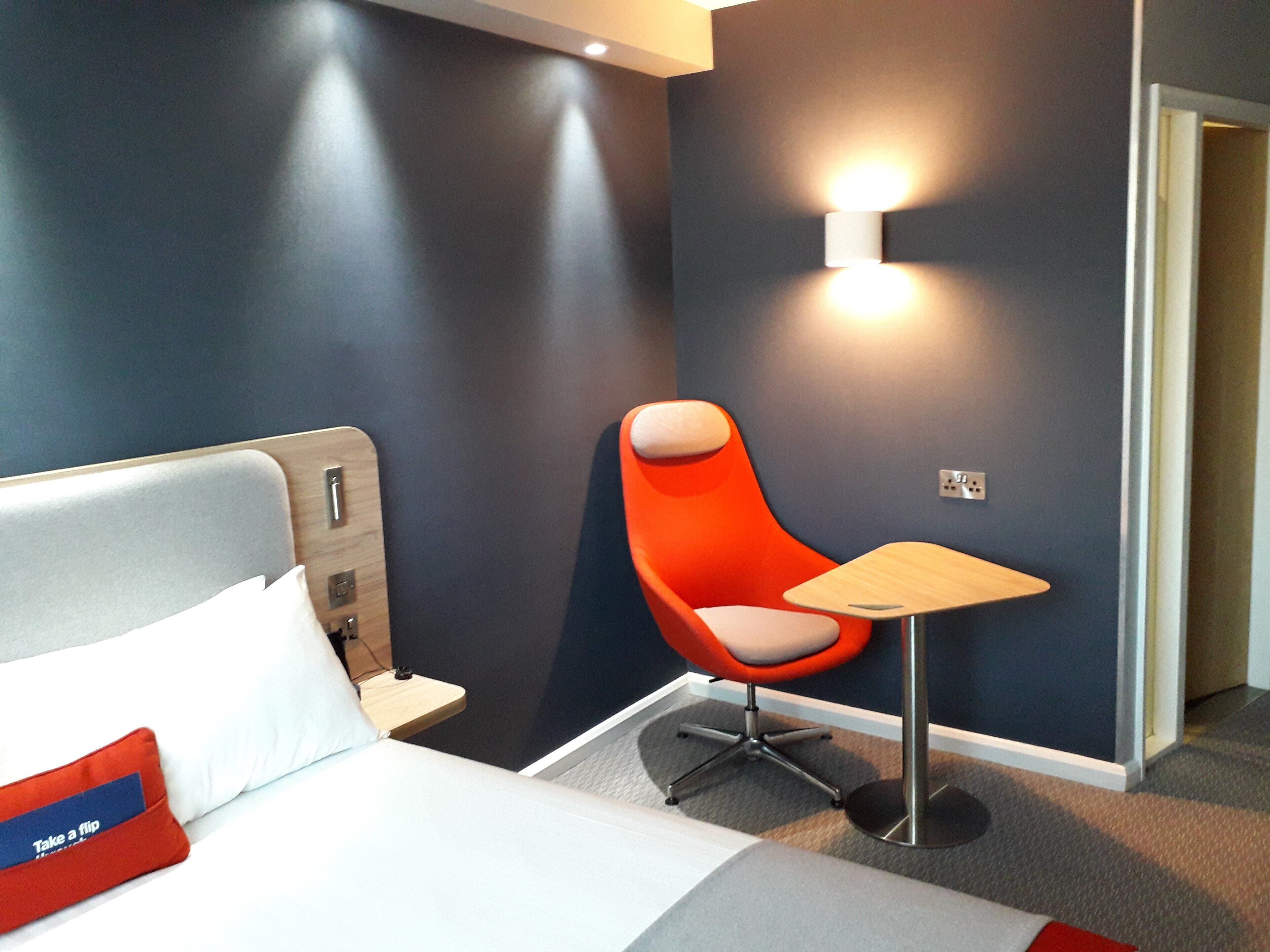 Une chambre d’hôtel avec un lit, une chaise rouge, une petite table et un lampadaire fixé au mur.