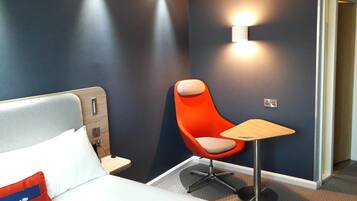 Une chambre d’hôtel avec un lit, une chaise rouge, une petite table et un lampadaire fixé au mur.