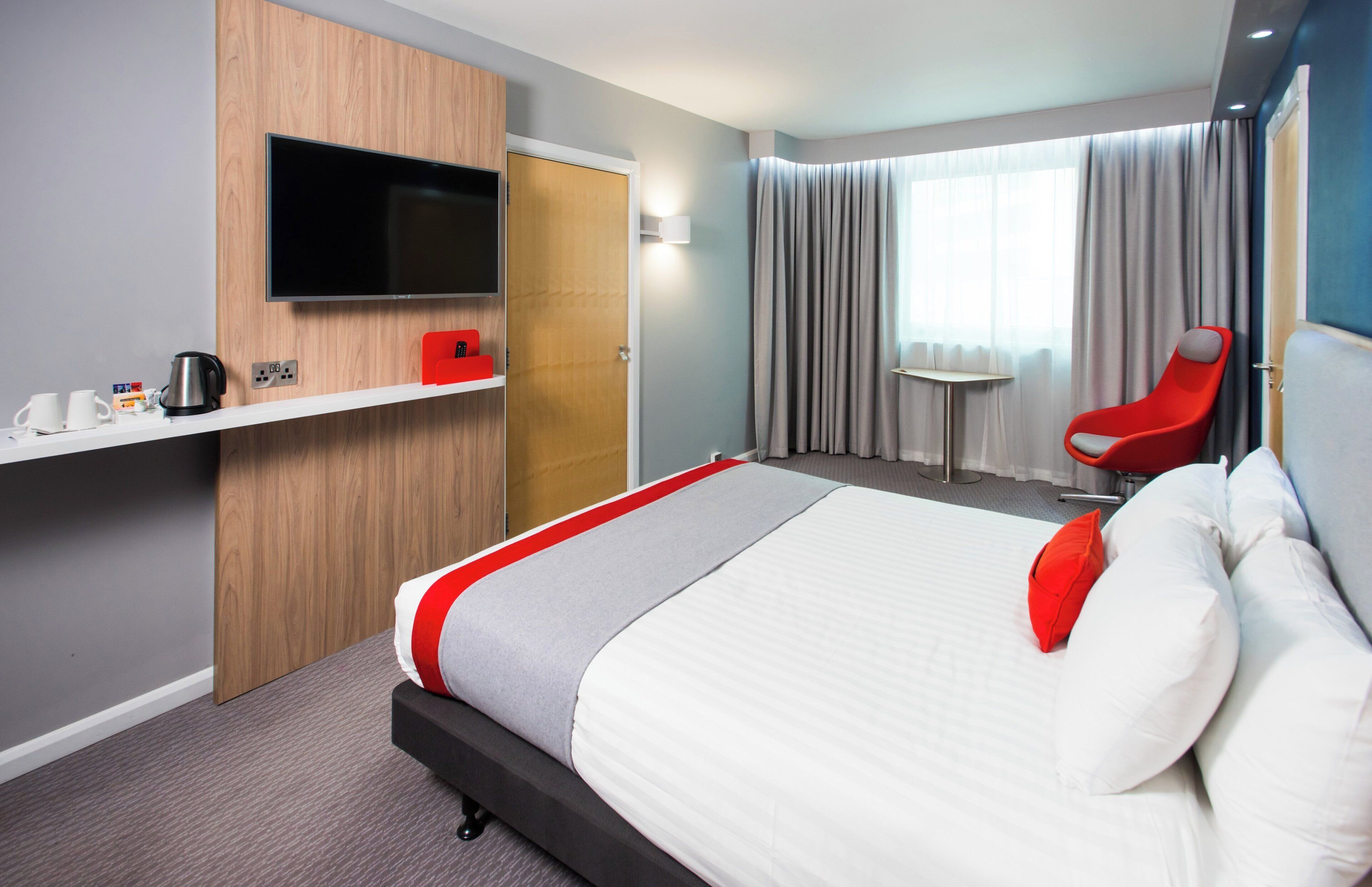 Une chambre d’hôtel comprenant un lit, une télévision, un bureau avec une bouilloire, une chaise et une fenêtre avec des rideaux.