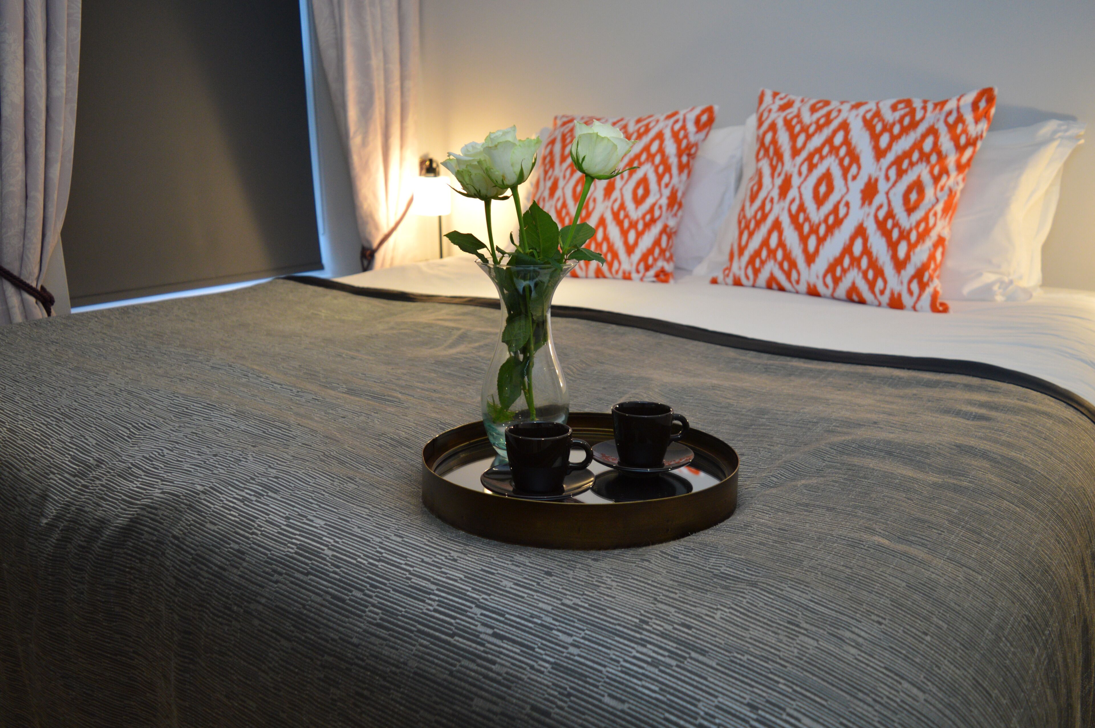 Un lit bien fait, sur lequel repose un plateau avec un vase contenant une fleur et deux tasses, le tout posé sur une couverture à motifs.