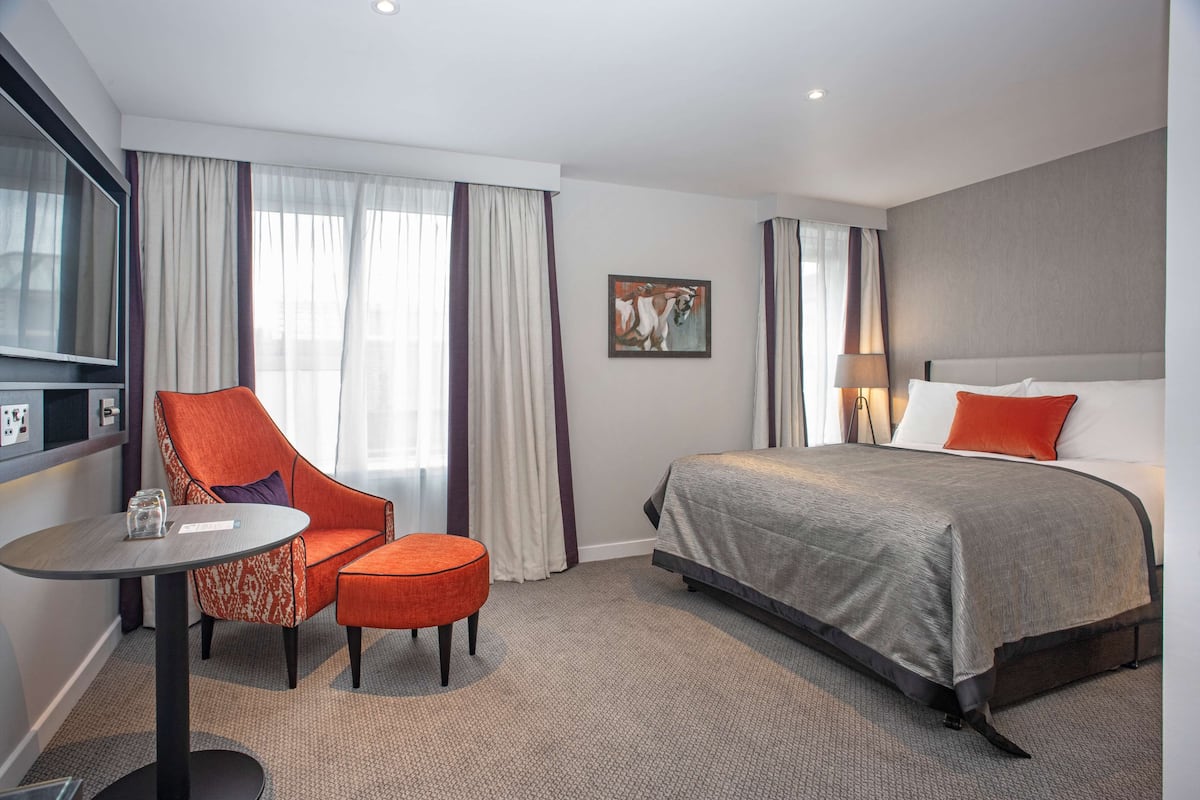 A hotel room with a bed, a chair, a table, and a painting on the wall.