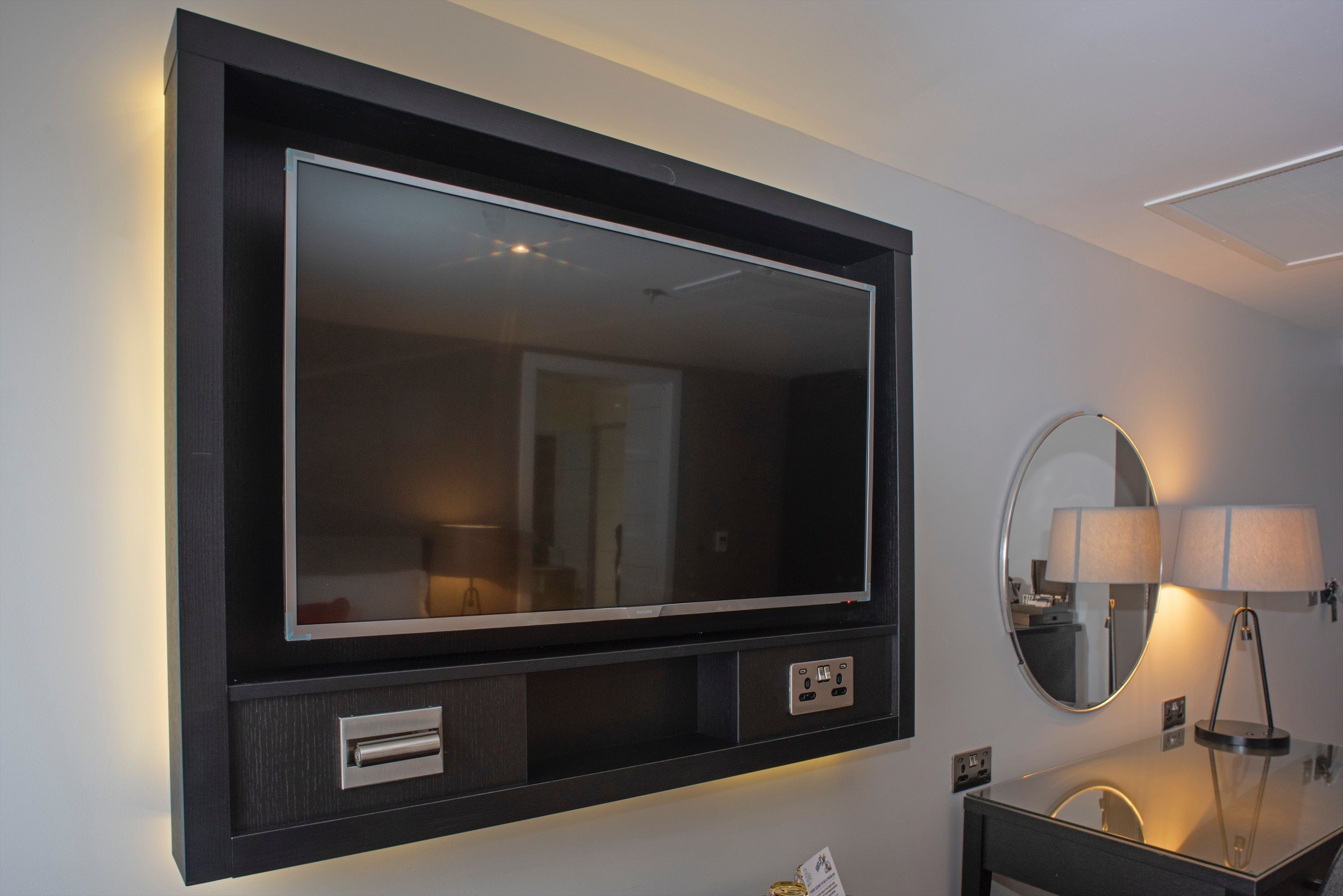 A modern hotel room with a large wall-mounted TV, a round mirror, and a bedside table with lamps.