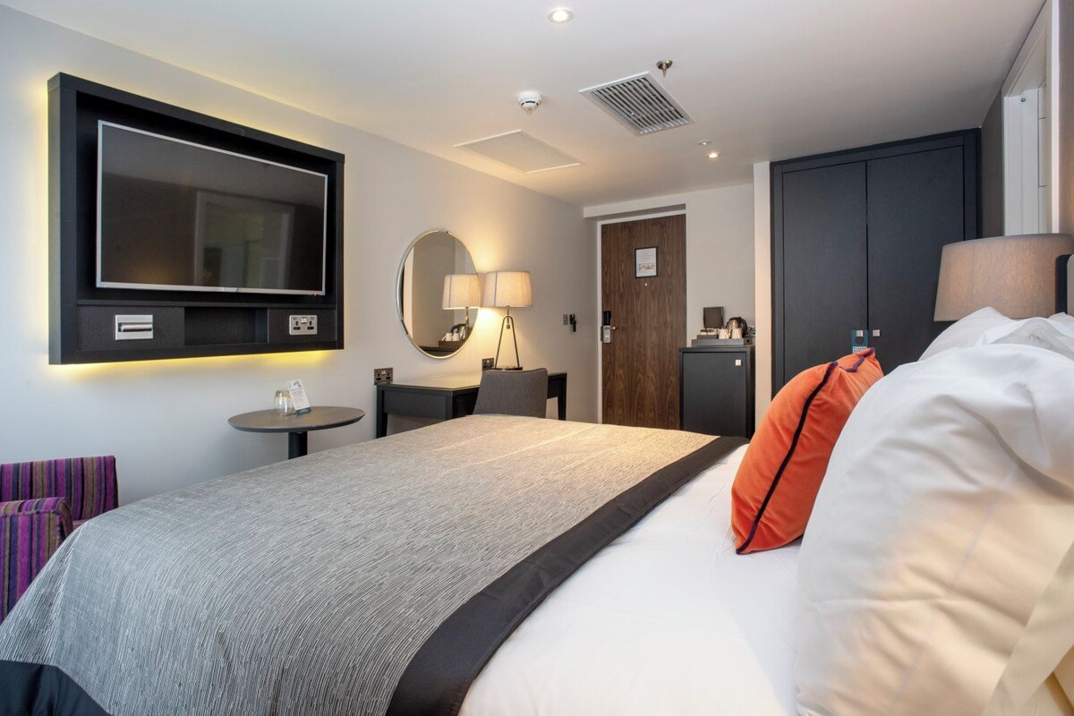 Quarto de hotel com cama, televisão na parede, escrivaninha com cadeira, luminária e guarda-roupa.