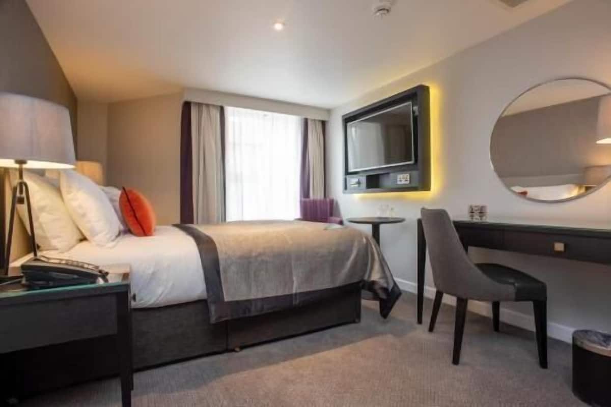 A hotel room with a bed, bedside table, desk, chair, TV, and a large mirror.
