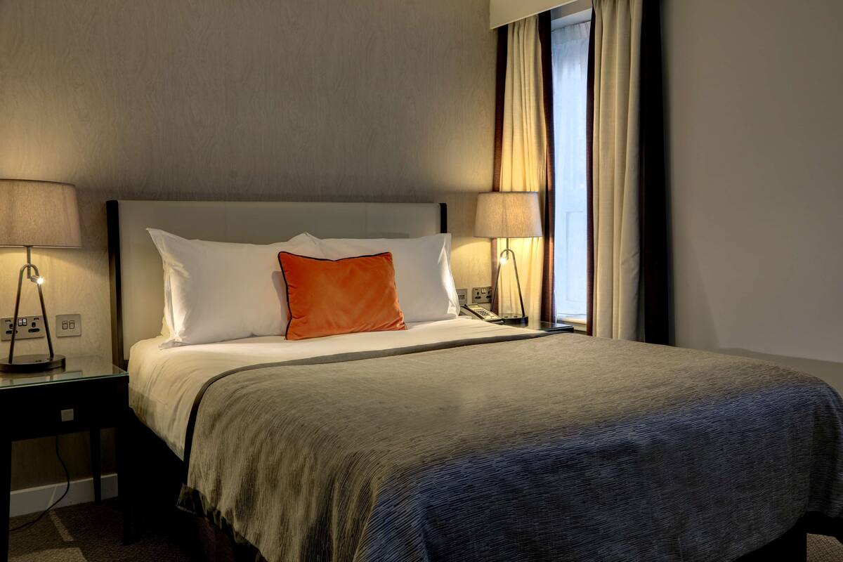 A hotel room with a bed, two bedside lamps, a nightstand, and a window with curtains.