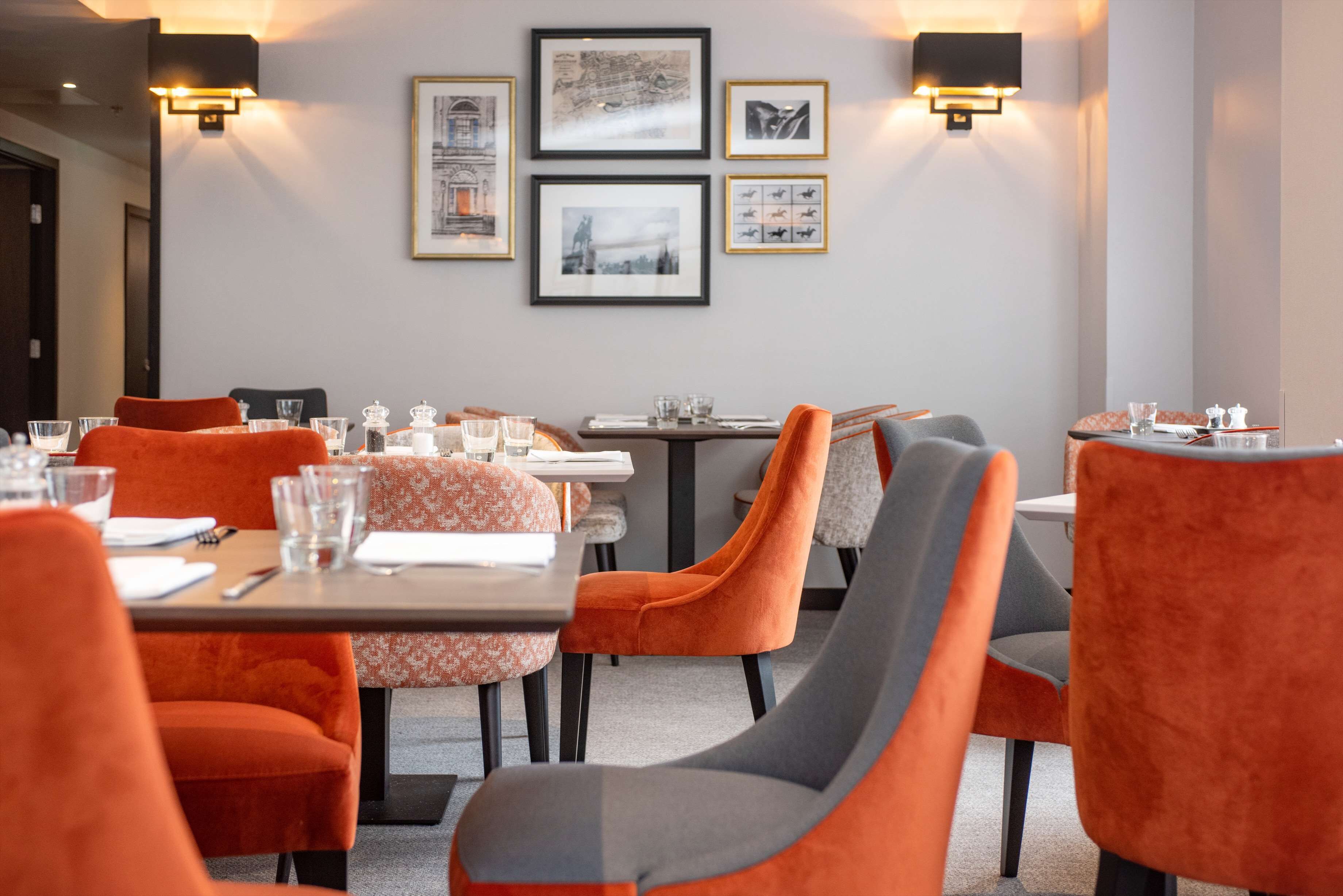 Uma sala de jantar com cadeiras estofadas em tons de laranja e cinza, mesas dispostas com copos e talheres, e quadros decorativos nas paredes.