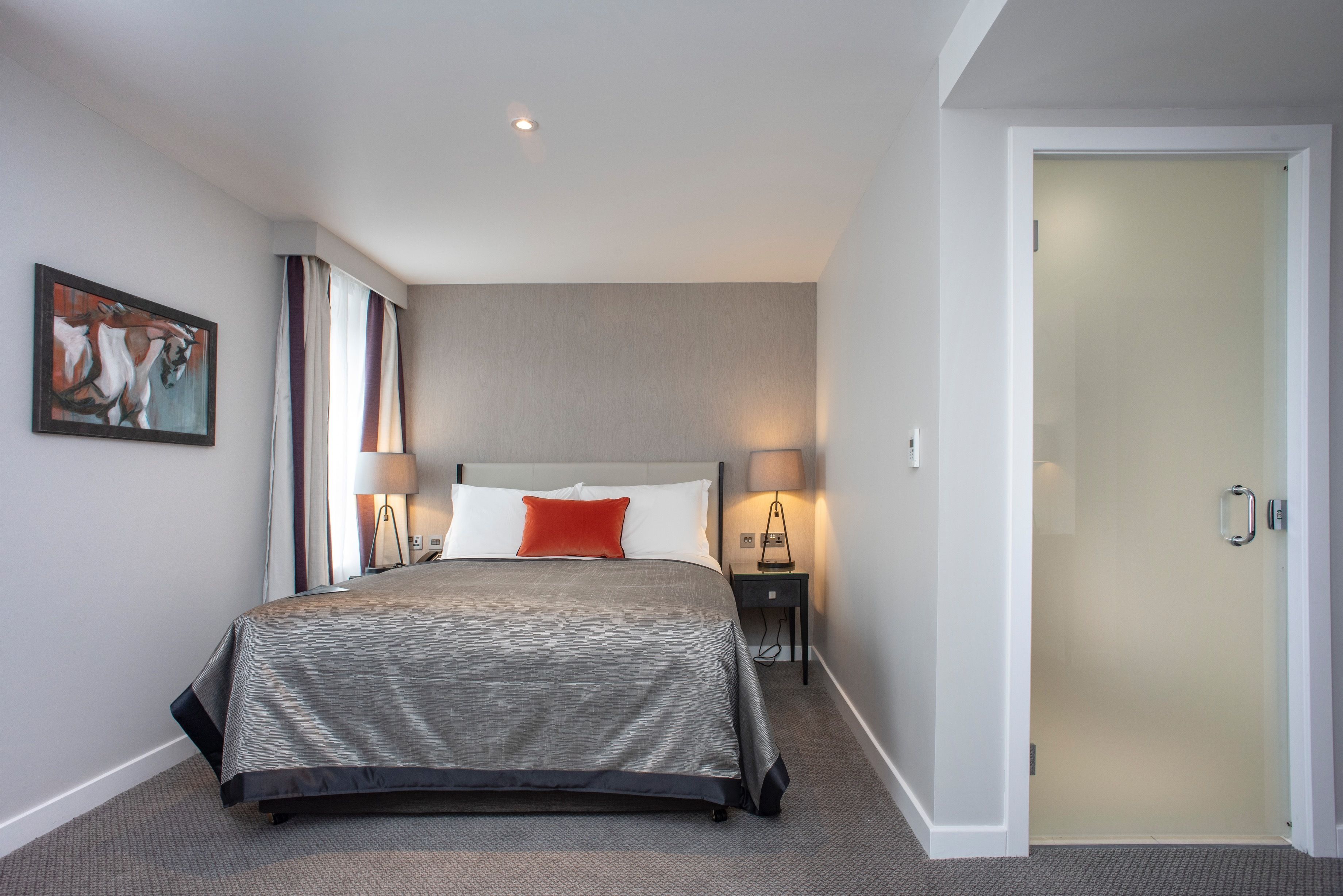 A modern hotel room with a large bed, two bedside tables with lamps, a painting on the wall, and a door to the right.