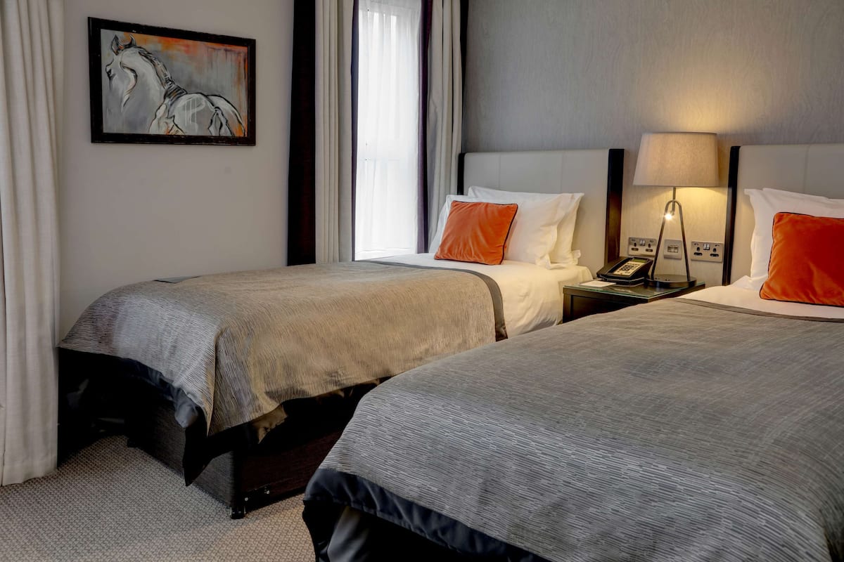 A hotel room with two beds, a painting on the wall, a nightstand with a lamp, and a window with curtains.