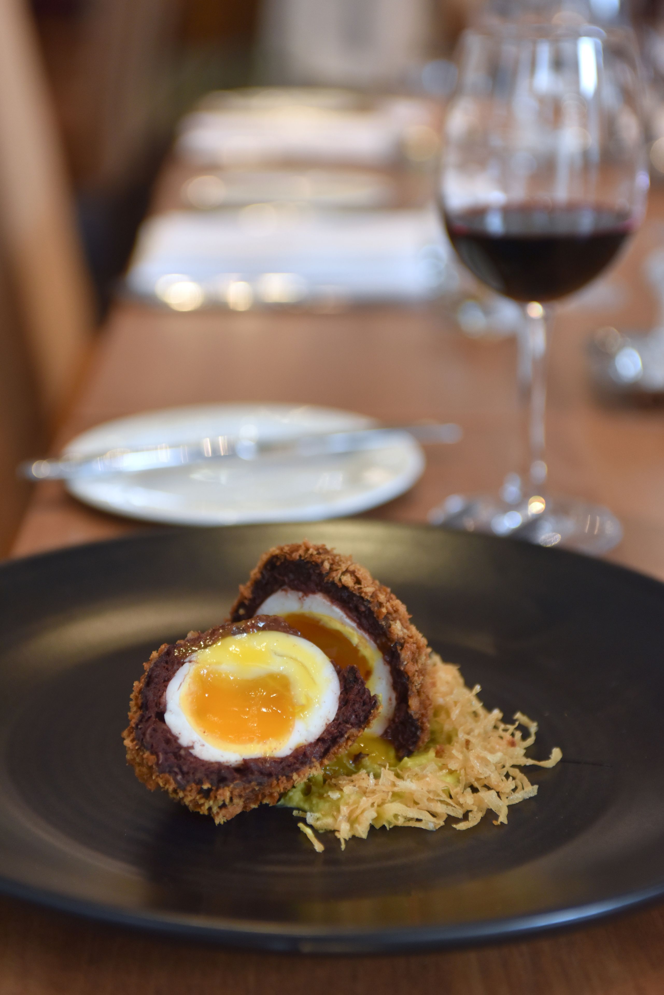 Un œuf écossais sur une assiette, avec un verre de vin rouge en arrière-plan.