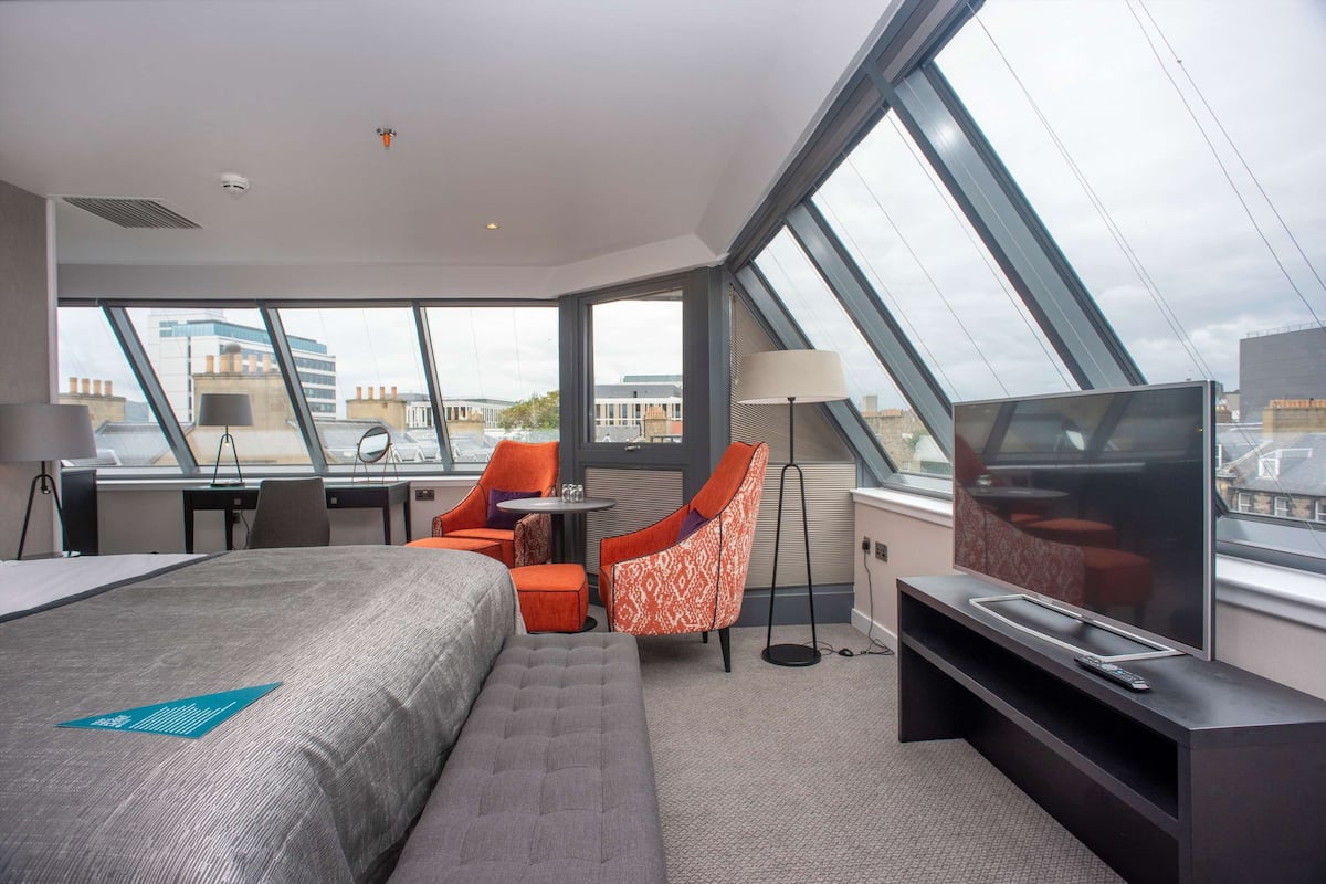 A modern hotel room with a large bed, a desk, a chair, a TV, and large windows offering city views.